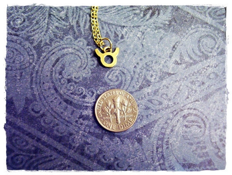 May include: Gold Taurus zodiac sign pendant necklace on a blue patterned background with a US dime for scale.