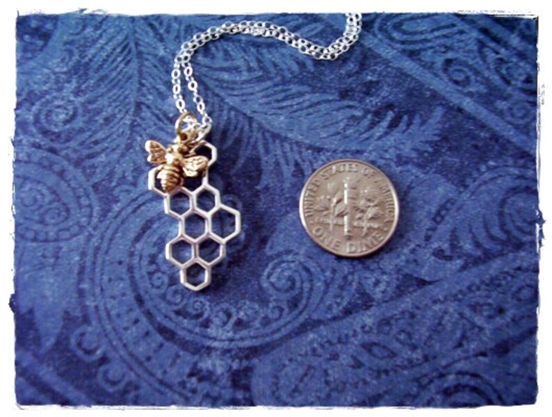 May include: A silver chain necklace with a silver honeycomb pendant and a gold bee charm. The necklace is on a blue patterned background.