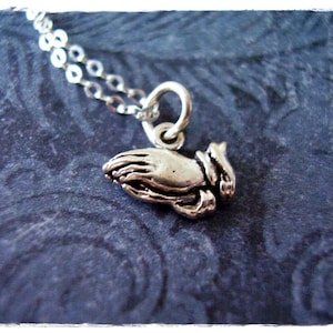 May include: A silver necklace with a praying hands charm. The charm is detailed and has a textured finish.