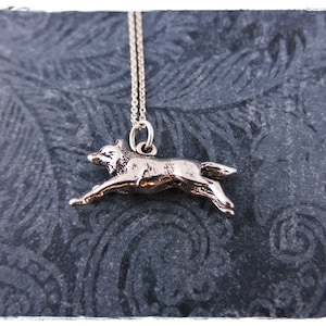 May include: A silver-colored pendant necklace featuring a running wolf. The wolf pendant is detailed and suspended from a delicate chain. The necklace is displayed against a dark, textured background.