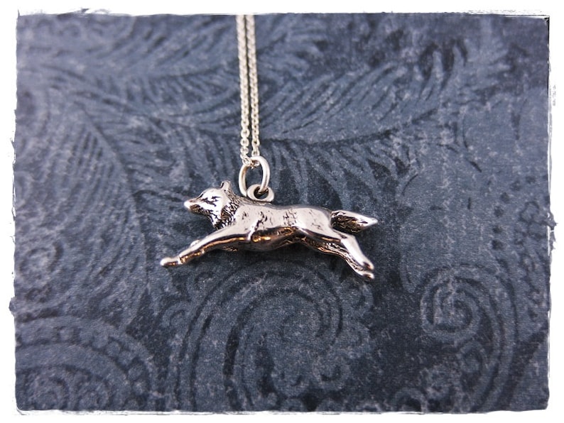 May include: A silver-colored pendant necklace featuring a running wolf. The wolf pendant is detailed and suspended from a delicate chain. The necklace is displayed against a dark, textured background.