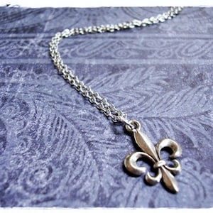 May include: A silver chain necklace with a silver fleur-de-lis pendant. The necklace is on a blue patterned background.