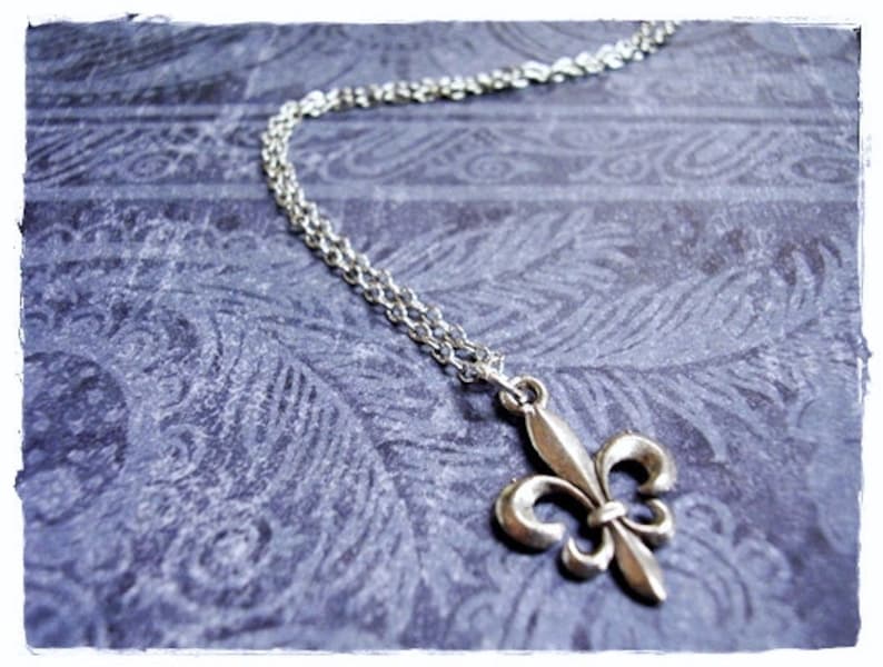 May include: A silver chain necklace with a silver fleur-de-lis pendant. The necklace is on a blue patterned background.