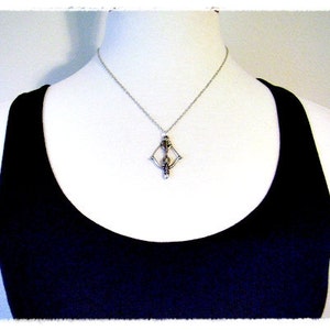 May include: A silver necklace with a geometric pendant featuring a stylized figure. The necklace is worn by a person wearing a black tank top.