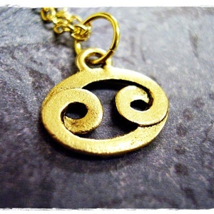 May include: Gold-toned pendant necklace featuring a stylized Cancer zodiac symbol.
