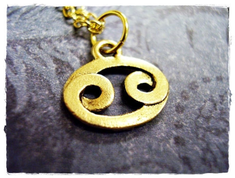 May include: Gold-toned pendant necklace featuring a stylized Cancer zodiac symbol.
