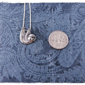 May include: A silver sloth pendant necklace hanging from a silver chain. The pendant is about the size of a UK ten pence coin.