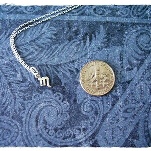 May include: A silver necklace with a small silver charm in the shape of the astrological sign Scorpio. The necklace is lying on a blue patterned fabric background. A US dime coin is also visible in the image.