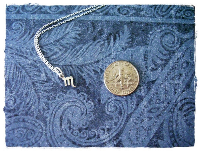 May include: A silver necklace with a small silver charm in the shape of the astrological sign Scorpio. The necklace is lying on a blue patterned fabric background. A US dime coin is also visible in the image.
