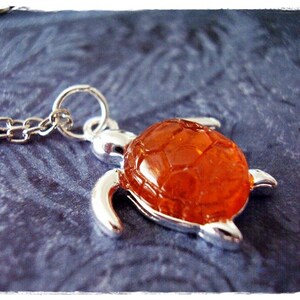 Könnte beinhalten: Eine silberne Schildkrötenkette mit einer orangefarbenen Glasschale. Die Schale hat ein strukturiertes Design.