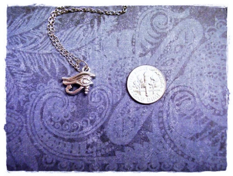 May include: Silver necklace with an eye of Horus pendant on a purple background with a paisley pattern.