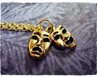 May include: A gold necklace with a pendant featuring two small, gold-colored masks. One mask is smiling and the other is frowning.