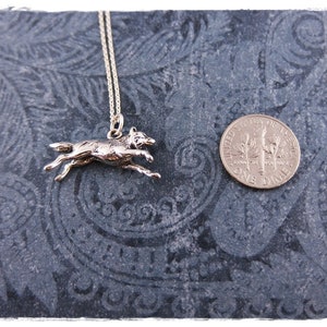 May include: A silver-colored pendant necklace featuring a running wolf charm. The wolf is detailed and appears to be in motion. A U.S. dime is next to the pendant for size comparison. The necklace is on a dark, patterned surface.