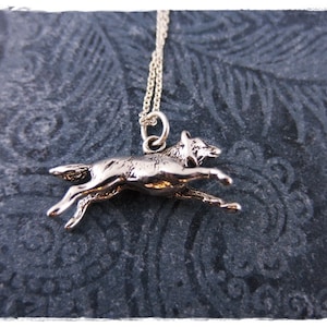 May include: A silver-colored pendant necklace featuring a running wolf charm. The wolf is depicted in mid-stride, with its legs extended. The necklace has a delicate chain and is set against a dark, patterned background.