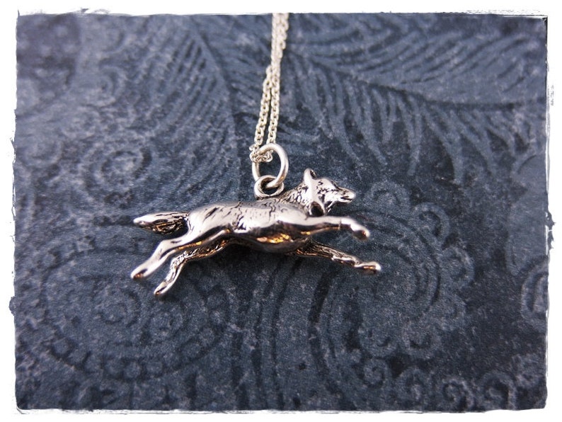 May include: A silver-colored pendant necklace featuring a running wolf charm. The wolf is depicted in mid-stride, with its legs extended. The necklace has a delicate chain and is set against a dark, patterned background.