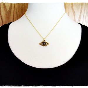 May include: A gold necklace with an eye pendant. The eye is dark brown and surrounded by a gold frame. The necklace is on a white mannequin.