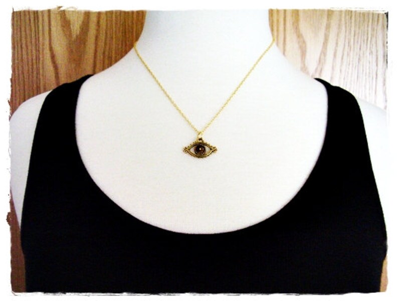 May include: A gold necklace with an eye pendant. The eye is dark brown and surrounded by a gold frame. The necklace is on a white mannequin.