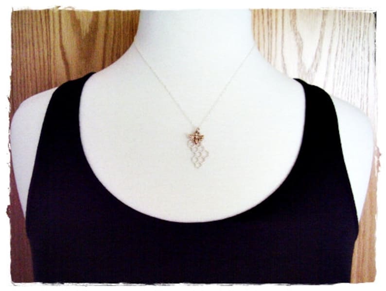 May include: A silver necklace with a honeycomb pendant and a small bee charm. The necklace is worn by a person wearing a black tank top.