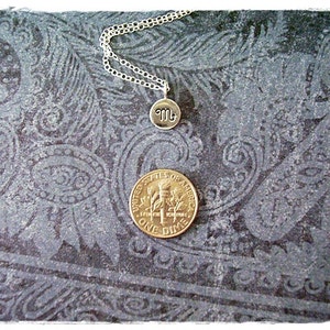 Pu&ograve; includere: Una collana d'argento con un piccolo ciondolo d'argento che presenta il simbolo astrologico dello Scorpione. La collana &egrave; esposta su uno sfondo blu scuro con un motivo paisley.