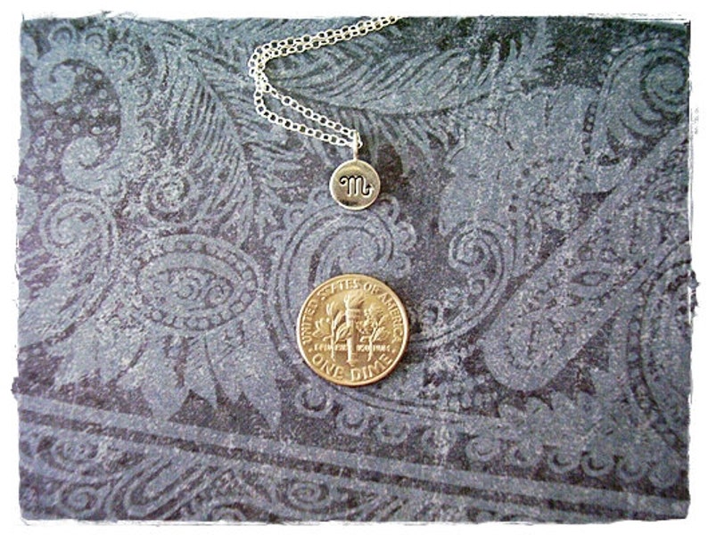 Pu&ograve; includere: Una collana d'argento con un piccolo ciondolo d'argento che presenta il simbolo astrologico dello Scorpione. La collana &egrave; esposta su uno sfondo blu scuro con un motivo paisley.
