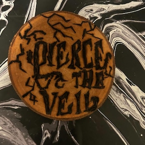 Puede incluir: Un posavasos redondo de madera con las palabras "PIERCE THE VEIL" quemadas en la madera en una fuente gótica. El posavasos tiene una veta de madera natural y está rodeado por un fondo arremolinado en blanco y negro.