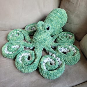 May include: A plush octopus toy crafted from variegated green yarn with a soft, fuzzy texture. The creature features a rounded head, two dark plastic eyes, and eight curled tentacles that reveal a white, textured underside with a speckled pattern.
