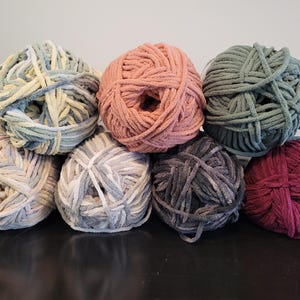 May include: A collection of eight yarn balls in various colors, including pink, green, gray, and purple. The yarn appears to be a soft, velvety texture, perfect for knitting or crocheting projects. The balls are arranged in a visually appealing display.