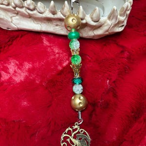 May include: A gold and green beaded clip with a gold dragon charm. The charm features a three-headed dragon with wings outstretched. The clip is on a red fuzzy surface. The text "THE GREENS" is visible in the bottom left corner of the image.