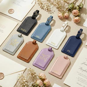 May include: A collection of rectangular luggage tags in black, blue, white, gray, tan, lavender, navy, and pink, each featuring a buckle strap and gold-stamped initials or icons, arranged alongside cream-colored wedding stationery and dried flowers.