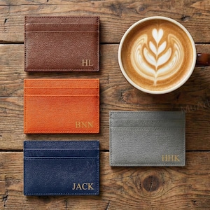 Puede incluir: Cuatro tarjeteros personalizados en marrón, naranja, azul marino y gris, cada uno con iniciales doradas. Una taza de café con arte latte está sobre una mesa de madera. Los tarjeteros son rectangulares con múltiples ranuras.