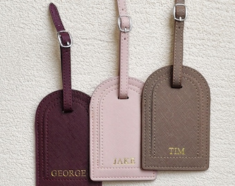 Può includere: Tre etichette per bagagli rettangolari con parte superiore arrotondata e cinturini regolabili con fibbia. Le etichette sono bordeaux scuro, rosa chiaro e marrone, con finitura testurizzata e nomi impressi in oro: GEORGE, JAKE e TIM.