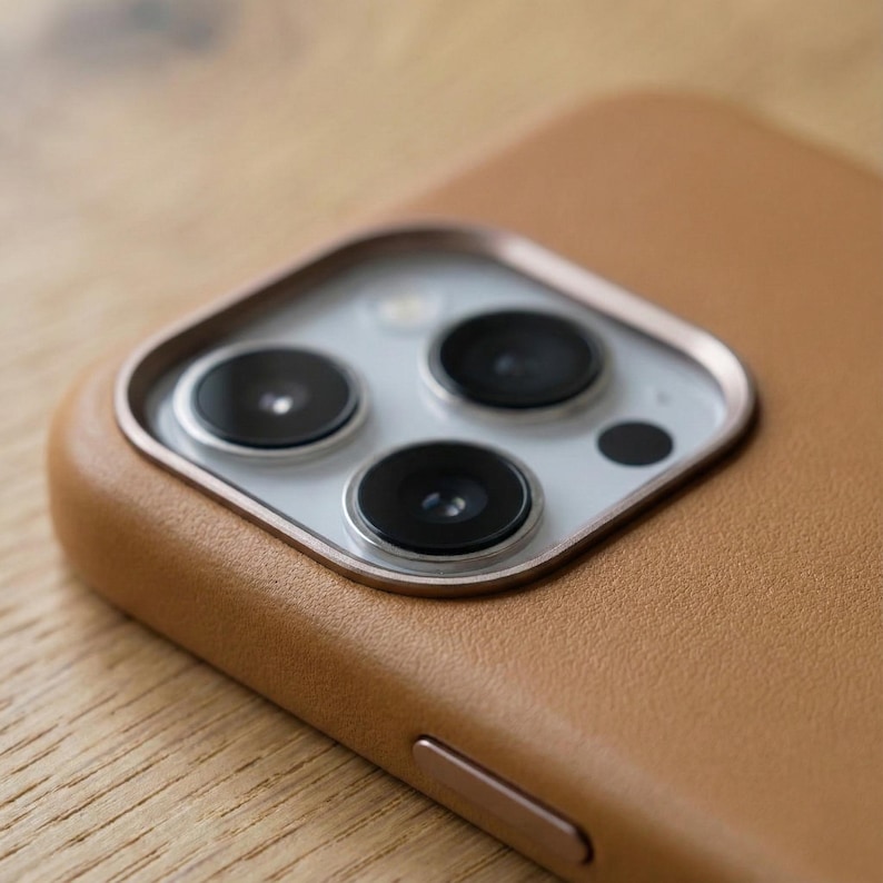 May include: Close-up of a tan leather phone case with a camera module. The case has a smooth texture and a metallic accent around the camera lenses. The camera module features three black lenses and a flash.