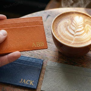 May include: An orange leather card holder with the initials "ABB" in gold lettering is held in a hand. Other card holders in navy blue and gray are also visible. A cup of coffee with latte art sits nearby.