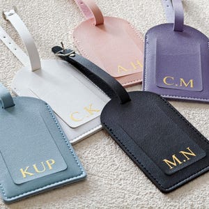 Puede incluir: Cinco etiquetas de equipaje rectangulares con correas ajustables en azul claro, blanco, rosa, morado y negro. Cada etiqueta presenta un borde cosido e iniciales doradas en relieve: KUP, C.K, A.H, C.M y M.N en la superficie frontal.