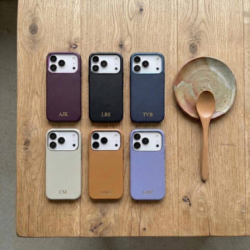 May include: A collection of six colorful phone cases in various shades, including purple, black, navy, beige, tan, and lavender. Each case has a camera cutout and a personalized monogram. A wooden spoon and a small plate are also visible.