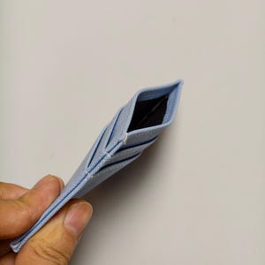 May include: A light blue card holder with multiple slots. The card holder is made of a textured fabric and has a black interior. The card holder is held in a hand against a white background.