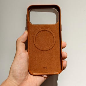May include: A brown leather phone case with a circular design on the back. The case has a rectangular camera cutout and the text "17PM" at the bottom. The case is held in a hand.