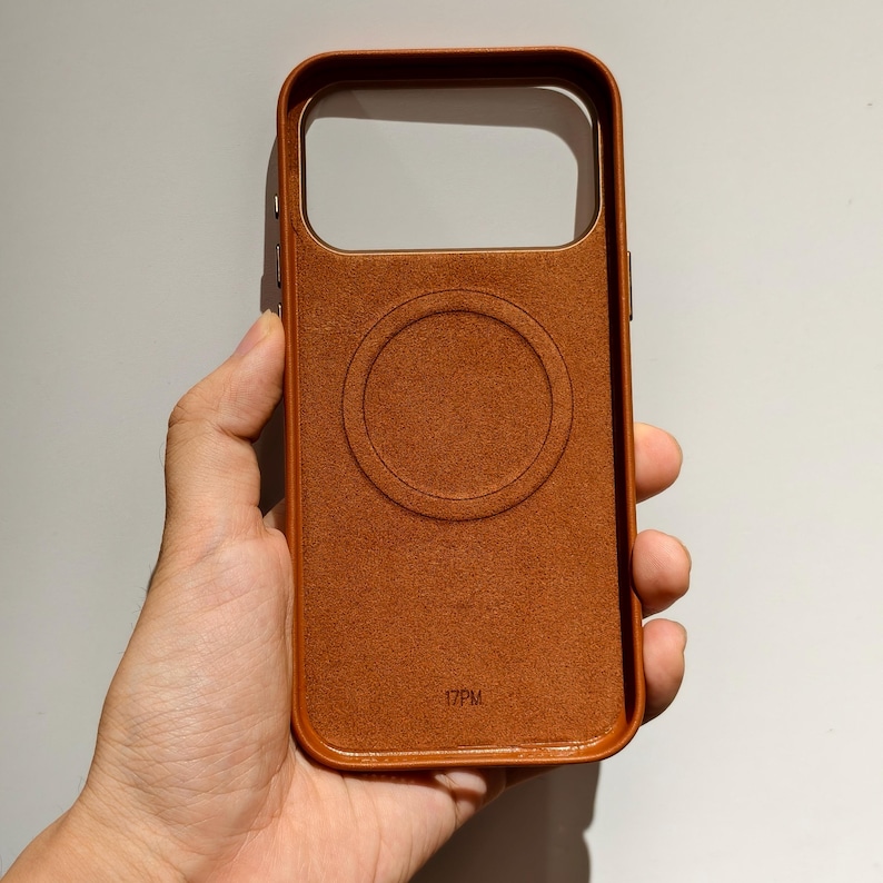 May include: A brown leather phone case with a circular design on the back. The case has a rectangular camera cutout and the text "17PM" at the bottom. The case is held in a hand.