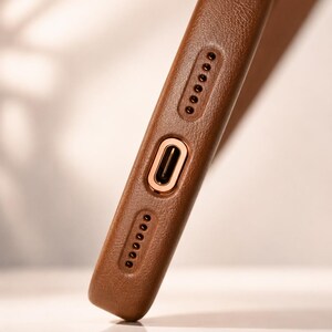May include: Close-up of a brown leather phone case. The case has a textured surface and features a gold-colored charging port and speaker holes. The case is designed to protect the phone.