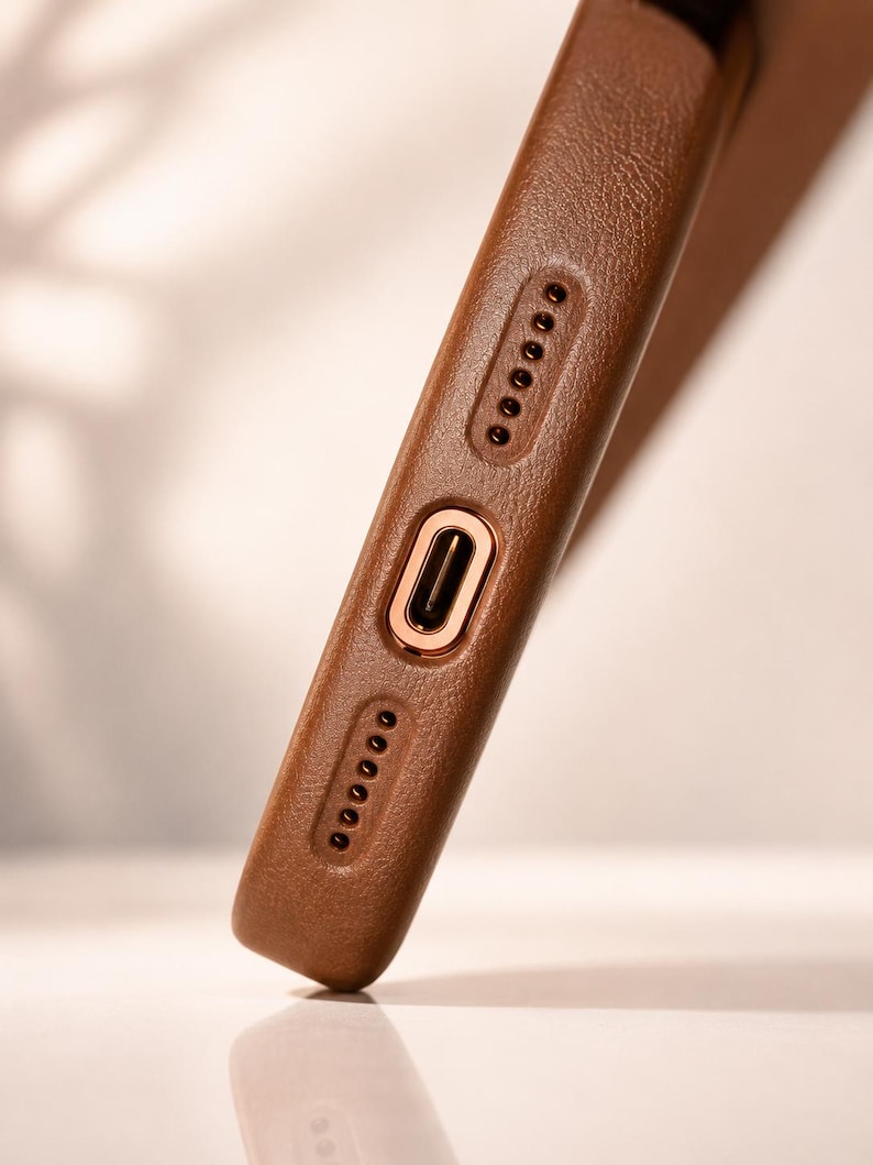 May include: Close-up of a brown leather phone case. The case has a textured surface and features a gold-colored charging port and speaker holes. The case is designed to protect the phone.
