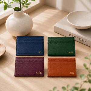 May include: Four leather cardholders in navy blue, green, burgundy, and orange. Each cardholder has a different set of initials in gold lettering: AK, HANK, KITE, and RD. The cardholders are arranged on a wooden surface.