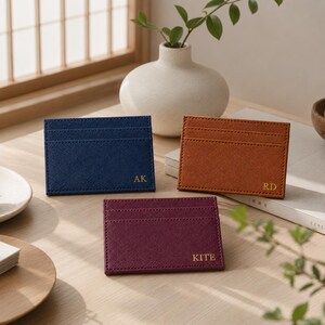 May include: Three leather cardholders in navy blue, brown, and burgundy are displayed on a wooden surface. Each cardholder has a different set of initials in gold lettering. A white vase with greenery is in the background.
