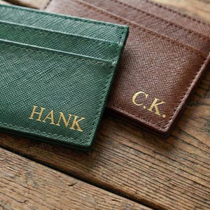 May include: Two personalized cardholders in green and brown, with gold lettering. The green cardholder is embossed with the name "HANK", while the brown one has the initials "C.K.". The cardholders are made of a textured material.