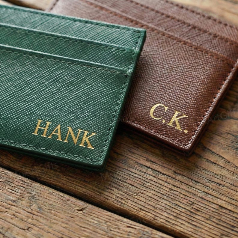 May include: Two personalized cardholders in green and brown, with gold lettering. The green cardholder is embossed with the name "HANK", while the brown one has the initials "C.K.". The cardholders are made of a textured material.