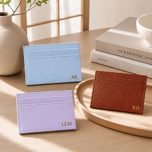 May include: Three personalized cardholders in pastel colors: light blue, lavender, and brown. Each cardholder features a stitched border and initials: "AK", "LLM", and "RD". The cardholders are displayed on a wooden tray.