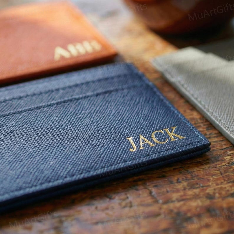 May include: A navy blue cardholder with the name "JACK" in gold lettering is in focus. Other cardholders in brown and gray are slightly blurred in the background. The cardholders are on a wooden surface.