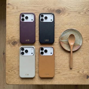 May include: Four personalized phone cases in plum, black, cream, and tan, each with initials. A small ceramic plate and wooden spoon are also on the wooden table. The cases have a camera cutout and a smooth finish.