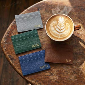 May include: Four personalized cardholders in gray, green, blue, and brown, each with gold lettering. A cup of coffee with latte art is next to the cardholders. The cardholders are on a wooden surface.