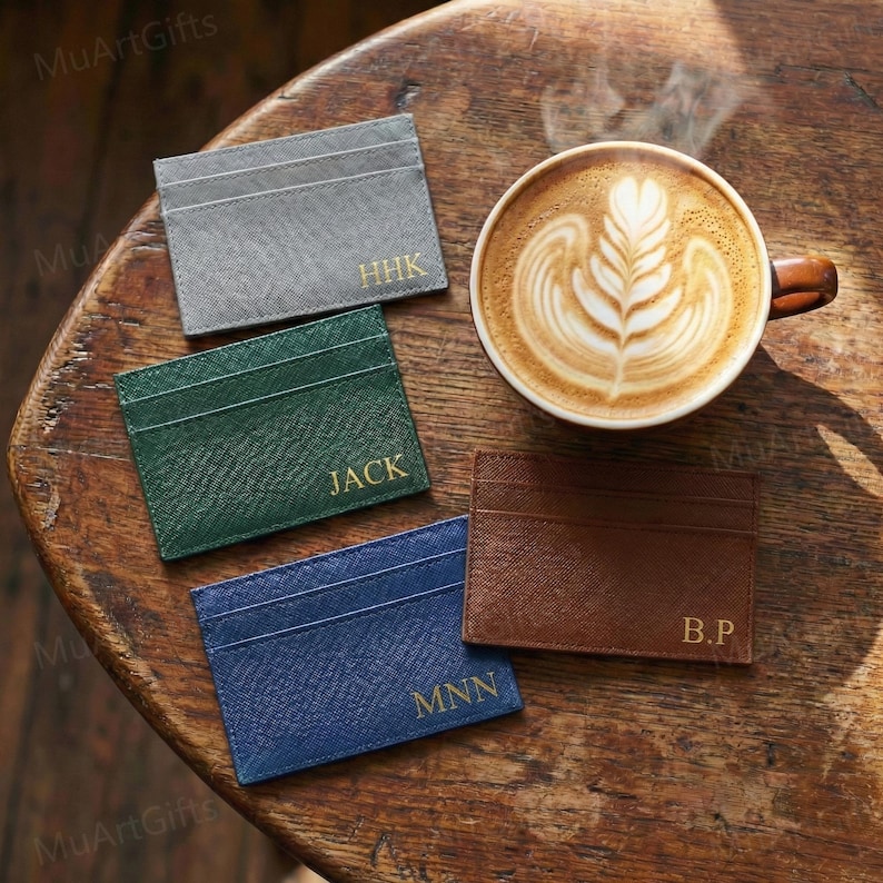 May include: Four personalized cardholders in gray, green, blue, and brown, each with gold lettering. A cup of coffee with latte art is next to the cardholders. The cardholders are on a wooden surface.