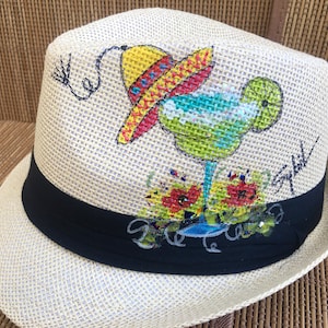 May include: A straw hat with a black band and a painted design of a margarita with a lime wedge, a sombrero, and flowers. The hat has the words "Be Cool" written on the band.
